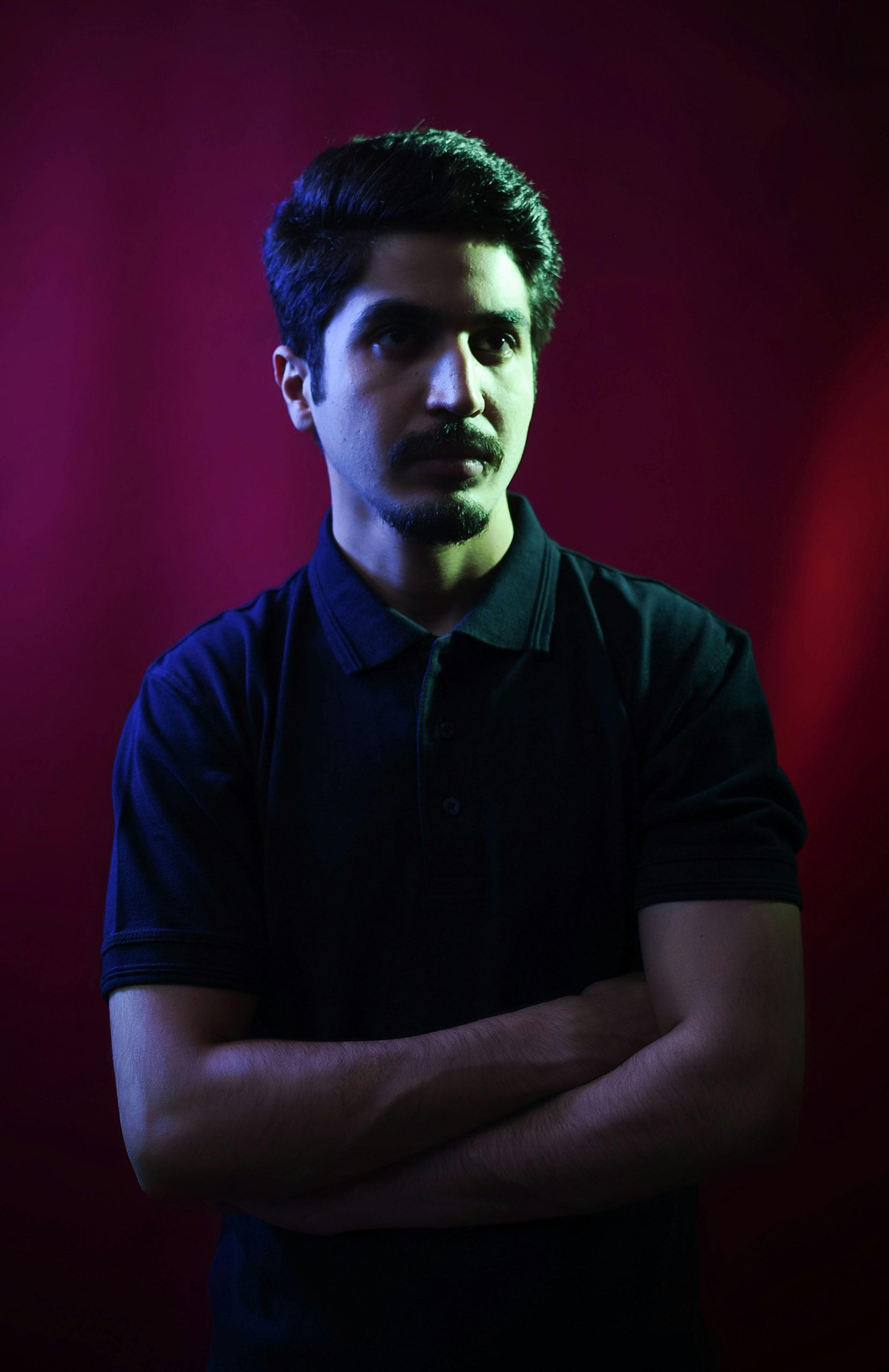 Moody portrait of a man with arms crossed in a darkly lit studio with red and blue lighting.