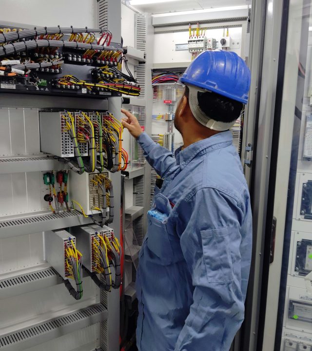 An electrician in a hard hat inspecting a control panel inside an industrial setting, ensuring safety and functionality.